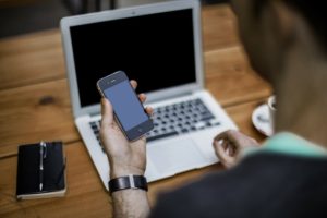 Image of a man holding an iPhone in his hand whilst facing a laptop.